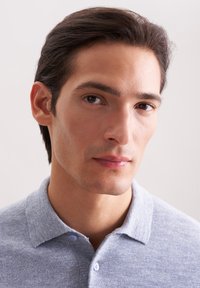 Junger Mann mit dunklem Haar und klarer Haut, der ein hellgraues, kurzärmliges Poloshirt trägt und leicht zur Seite schaut, vor einem schlichten Hintergrund.