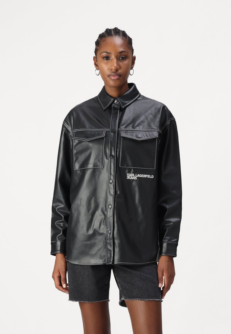 Giacca camicia nera in ecopelle con due tasche frontali, dettagli in cucitura bianca e logo "KARL LAGERFELD JEANS" sul lato sinistro in basso.