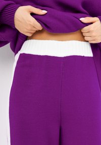 Pantalons en tricot violet avec une large ceinture côtelée blanche. Présente une texture lisse et des panneaux latéraux blancs, créant un effet de blocs de couleur.