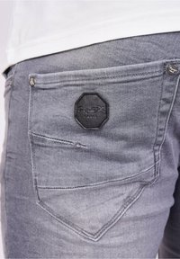Jeans in denim grigio con una finitura sbiadita, dotati di una toppa ottagonale nera sulla tasca posteriore e dettagli in metallo.