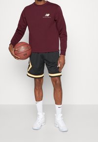 Hombre con sudadera de baloncesto maroon de New Balance, pantalones cortos de baloncesto negros y amarillos, calcetines blancos de Nike, zapatillas blancas, sosteniendo un balón de baloncesto Spalding.