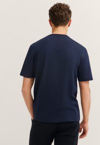 Mężczyzna w gładkiej, granatowej koszulce z krótkim rękawem i czarnych spodniach, stojący z rękami swobodnie wzdłuż ciała, zwrócony plecami do aparatu.