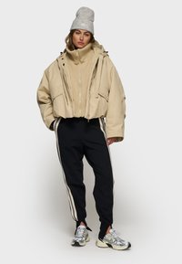 Beige oversized jakke med to forlommer, parret med sorte træningsbukser med hvide sidelister og sølv sneakers. Grå hue.