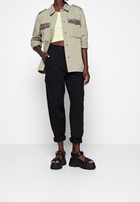 Light olive green button-up shirt with patterned pockets, paired with black cargo pants and chunky black sandals. Overall relaxed fit.