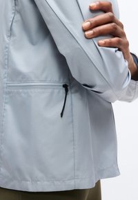 Άτομο διπλώνοντας το αριστερό χέρι πάνω από το στήθος, φοράει ανοιχτό γκρι μπουφάν με φερμουάρ σε πλαϊνή τσέπη και φαίνονται ελιάς πράσινο παντελόνι κάτω από το τελείωμα.