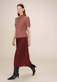 M pull à manches courtes tricoté en rose doux, associé à une jupe plissée bordeaux fluide et des bottes noires à talons pointus jusqu'aux genoux.