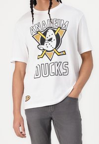 Persona vistiendo una camiseta blanca de los Anaheim Ducks con un logo de máscara de portero y dos palos de hockey cruzados en amarillo, combinada con pantalones grises.
