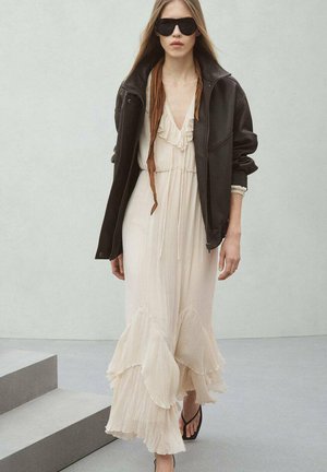 Donna che indossa un abito lungo beige con volant, una giacca nera oversize, grandi occhiali da sole neri e sandali neri, camminando all'interno su un pavimento grigio.