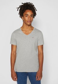 Camiseta de algodón gris con cuello en V y mangas cortas, con un pequeño logotipo bordado en el pecho. Textura suave y ajuste casual.