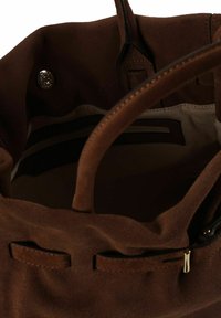 Intérieur d'un sac à main en daim marron avec une doublure lisse, deux poignées et une poche zippée ; doté d'une fermeture à pression et d'un logo métallique en accent.