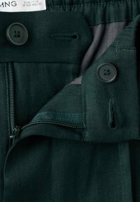 Pantalon vert foncé avec fermeture éclair partiellement ouverte et deux boutons noirs près de la taille, montrant la texture du tissu et les détails des coutures.