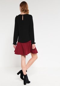 Černý top s dlouhým rukávem a průstřihem na zádech, doplněný rozšířenou vínovou sukní. Modelka má na sobě černé kotníkové boty.