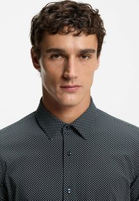 Svart skjorta med knappar och vita prickar, med spetsig krage och slät textur, designad för en figurnära passform.