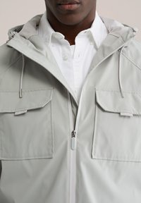 Lichtgrijze waterdichte jas met ritssluiting, twee voorzakken en een met mesh gevoerde capuchon met verstelbare trekkoorden.