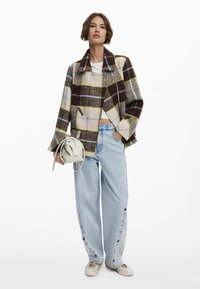 Kariertes Jackett in Braun-, Creme- und Grüntönen; weite blaue Jeans mit Knopfdetails; kleine Handtasche in Creme; legere Schuhe.