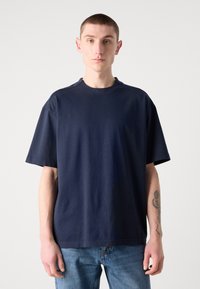 Námorně modré bavlněné tričko s krátkým rukávem, volný střih, kulatý výstřih a diskrétní detail loga značky blízko límce.