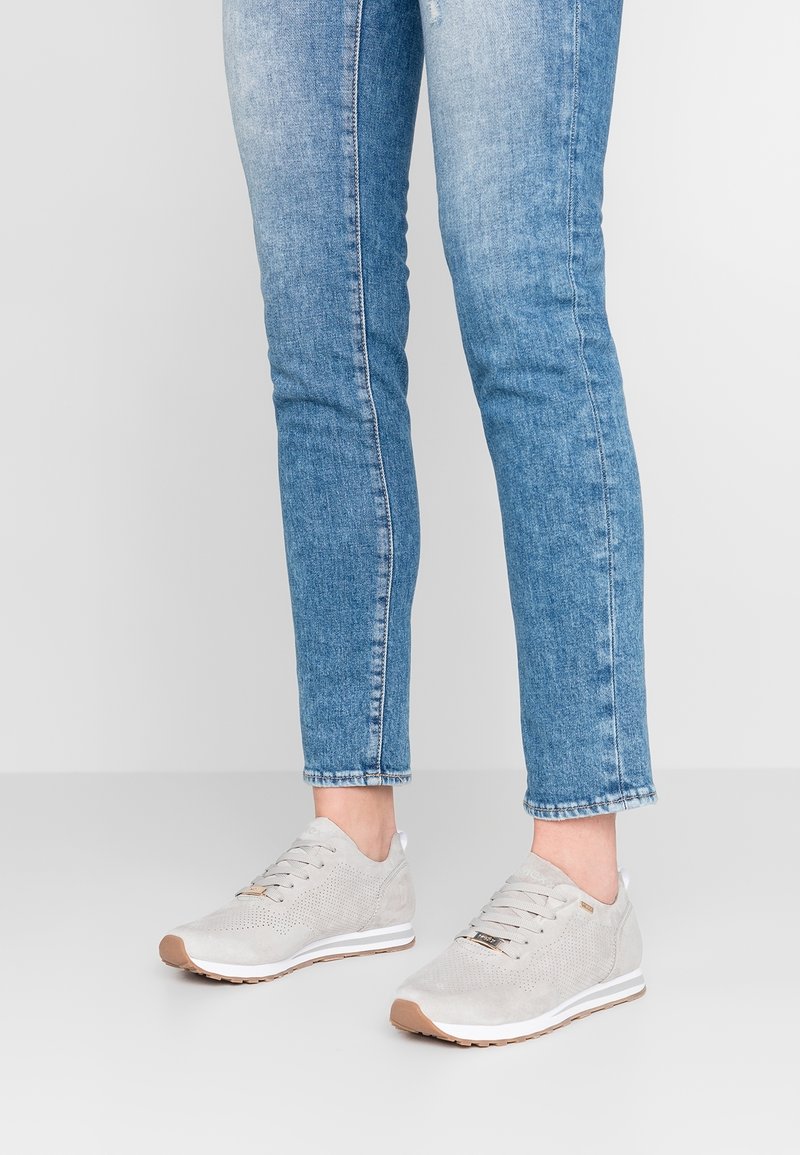 Personne portant des baskets à lacets gris clair avec des semelles intermédiaires blanches et un jean slim bleu sur un fond uni clair.