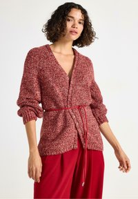 Kvinde iført en rød og hvid strikket cardigan med opsmøgte ærmer, snøret ind i taljen med et tyndt rødt bælte, kombineret med røde brede bukser.