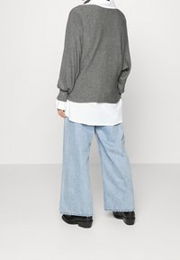 Grijze geribbelde trui met afgezakte schouders, gedragen over een wit overhemd met knopen, gecombineerd met lichtblauwe jeans met wijde pijpen en zwarte laarzen.