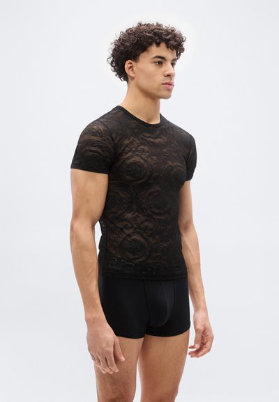 Νέος άνδρας που φοράει ένα διαφανές μαύρο δαντελένιο κοντομάνικο πουκάμισο και μαύρα σφιχτά εσώρουχα boxer, στέκεται μπροστά σε ένα απλό ανοιχτόχρωμο φόντο.