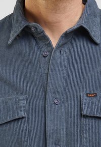 Camicia in corduroy blu scuro con superficie strutturata, dotata di colletto, chiusura frontale con bottoni, tasca e etichetta "Lee" arancione sulla tasca del petto.