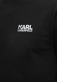 Μαύρο ύφασμα με λευκό κείμενο λογότυπου "KARL LAGERFELD", με μια μικρή σιλουέτα του κεφαλιού του Karl Lagerfeld ενσωματωμένη στο γράμμα "K".