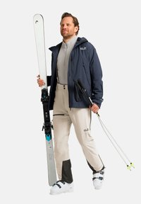 Skiuitrusting omvat witte ski's, beige skibroek met donkere accenten, en een marineblauwe jas. De skier houdt skistokken vast en draagt witte skischoenen.