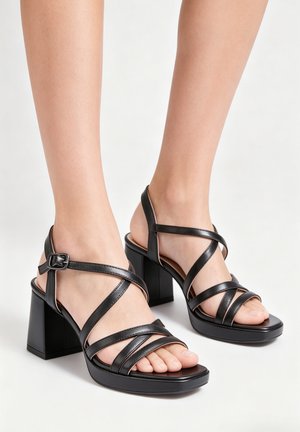 Pieds portant des sandales en cuir noir à talons épais avec plusieurs fines brides et une fermeture à boucle autour de la cheville sur fond blanc.