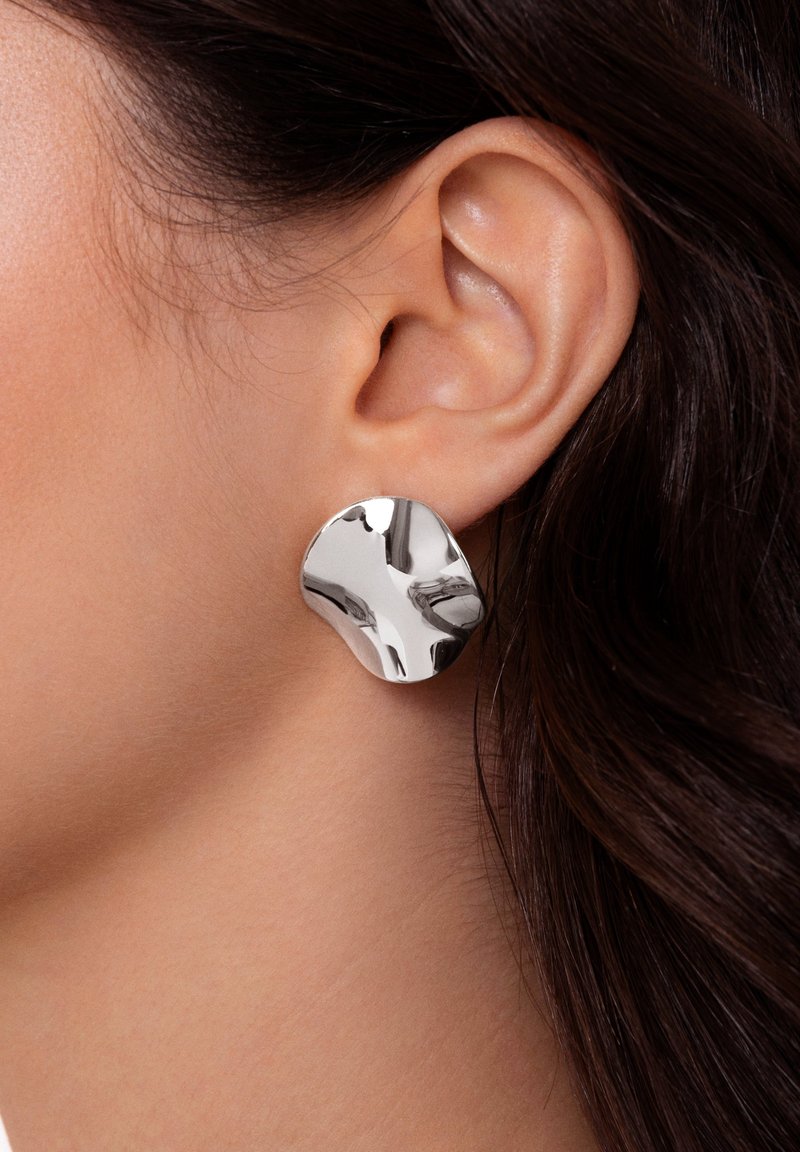 Gros plan d'une oreille avec une personne portant une boucle d'oreille en argent brillant de forme irrégulière, cheveux châtain foncé visibles en arrière-plan.
