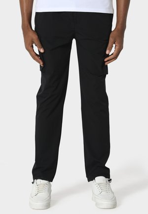 Person iført sorte cargobukser og hvide sneakers, stående med hænder afslappet langs siderne mod en ensartet lys baggrund.