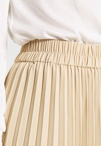 Falda beige con pliegues y cintura elástica, hecha de una tela suave, que muestra una textura vertical y un sutil brillo; combinada con una blusa clara.