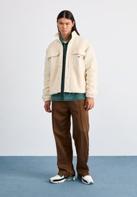 Giacca in shearling beige e tessuto con bottoni a pressione e due tasche sul petto, abbinata a una camicia verde, pantaloni marroni e sneakers multicolori.