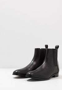 Bottes Chelsea en cuir noir avec panneaux latéraux élastiques, bout arrondi et talon bas et empilé. Texture lisse et design minimaliste.