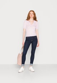Polo de manga curta listrado em roxo claro, jeans ajustados escuros e sapatilhas brancas. Segurando uma bolsa tote em rosa claro, em pé contra um fundo simples.