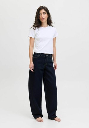 Woman with long dark hair stands barefoot wearing a white short-sleeve shirt and loose dark blue denim jeans against a plain background.
