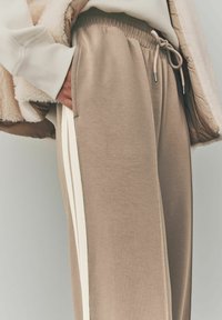 Pantalons de survêtement beiges avec une taille à cordon, dotés de poches latérales et de rayures verticales crème le long des coutures extérieures, fabriqués en tissu doux.