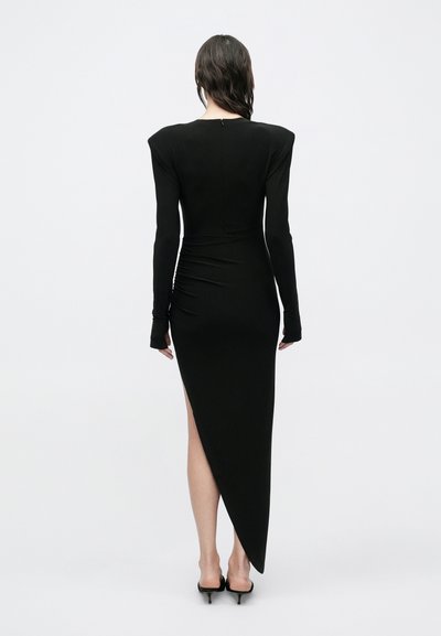 Μαύρο εφαρμοστό φόρεμα με μακριά μανίκια, επωμίδες, ασύμμετρη γραμμή στο τελείωμα και ρυτιδωτή υφή στο πίσω μέρος, σε συνδυασμό με ψηλοτάκουνα παπούτσια.