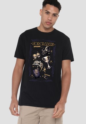 Junger Mann trägt ein schwarzes T-Shirt mit goldenen Vintage-Illustrationen magischer Objekte, die mit "Horcruxes" in einem stilisierten Rahmen bezeichnet sind.