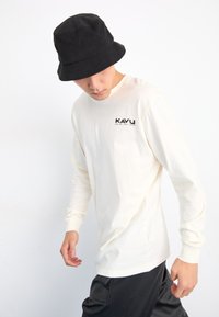 Maglietta a maniche lunghe in cotone crema con logo "KAVU" nero sul lato sinistro del petto, abbinata a un cappello a secchiello nero. Il modello indossa pantaloni neri.
