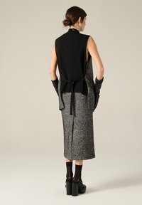 Top noir sans manches avec un col haut et un lien dans le dos, associé à une jupe grise à motifs et des gants noirs, sur fond neutre.