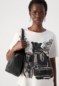 Vrouw in een wit T-shirt met een zwart panterafbeelding, met een zwarte leren handtas over haar schouder tegen een effen achtergrond.