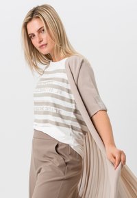 Un cardigan beige sur une chemise blanche avec des rayures beiges, présentant un léger éclat sur les manches. Un pantalon beige sur mesure avec une texture douce.