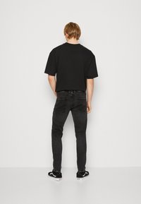 Ung man som står vänd mot en vit vägg, klädd i en svart t-shirt, mörka smala jeans och svarta sneakers med vita sulor.