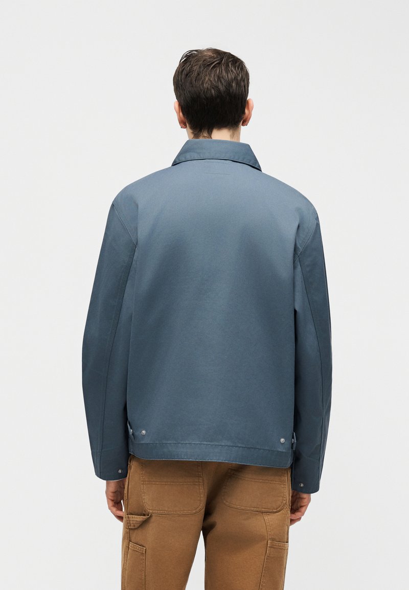 Uomo con giacca blu con colletto e pantaloni marroni di spalle su uno sfondo bianco uniforme.