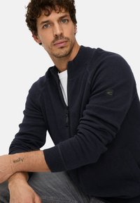 Navy-blue zip-up cardigan in textured knit, featuring a high collar, zippered front, and logo detail on the left sleeve.