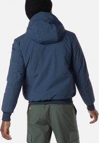 Giacca a vento blu navy con cappuccio, dotata di polsini e orlo a costine; indossata sopra pantaloni cargo verdi. Tessuto liscio e design isolato.