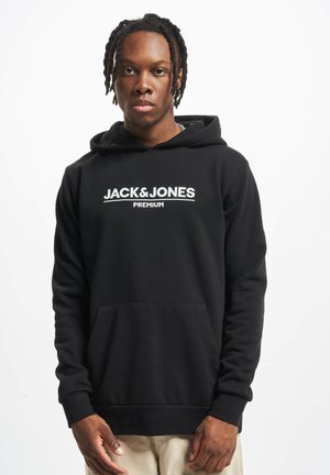 Joven con el cabello trenzado, vistiendo una sudadera negra con el logotipo "JACK & JONES PREMIUM", de pie contra un fondo blanco liso.