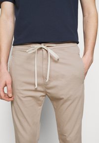 Beige bomullsblandade byxor med elastisk midja och krämfärgad snörning, med sidofickor och en slät, otexturerad yta.