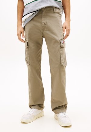 Άτομο που φοράει μπεζ cargo παντελόνια με τσέπες στα γόνατα και λευκά αθλητικά παπούτσια, στέκεται μπροστά σε απλό φόντο.