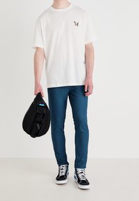 Biała bawełniana koszulka z haftem motyla na piersi, w połączeniu z turkusowymi spodniami. Czarna torebka o fakturze trzymana w jednej ręce; czarno-białe trampki.